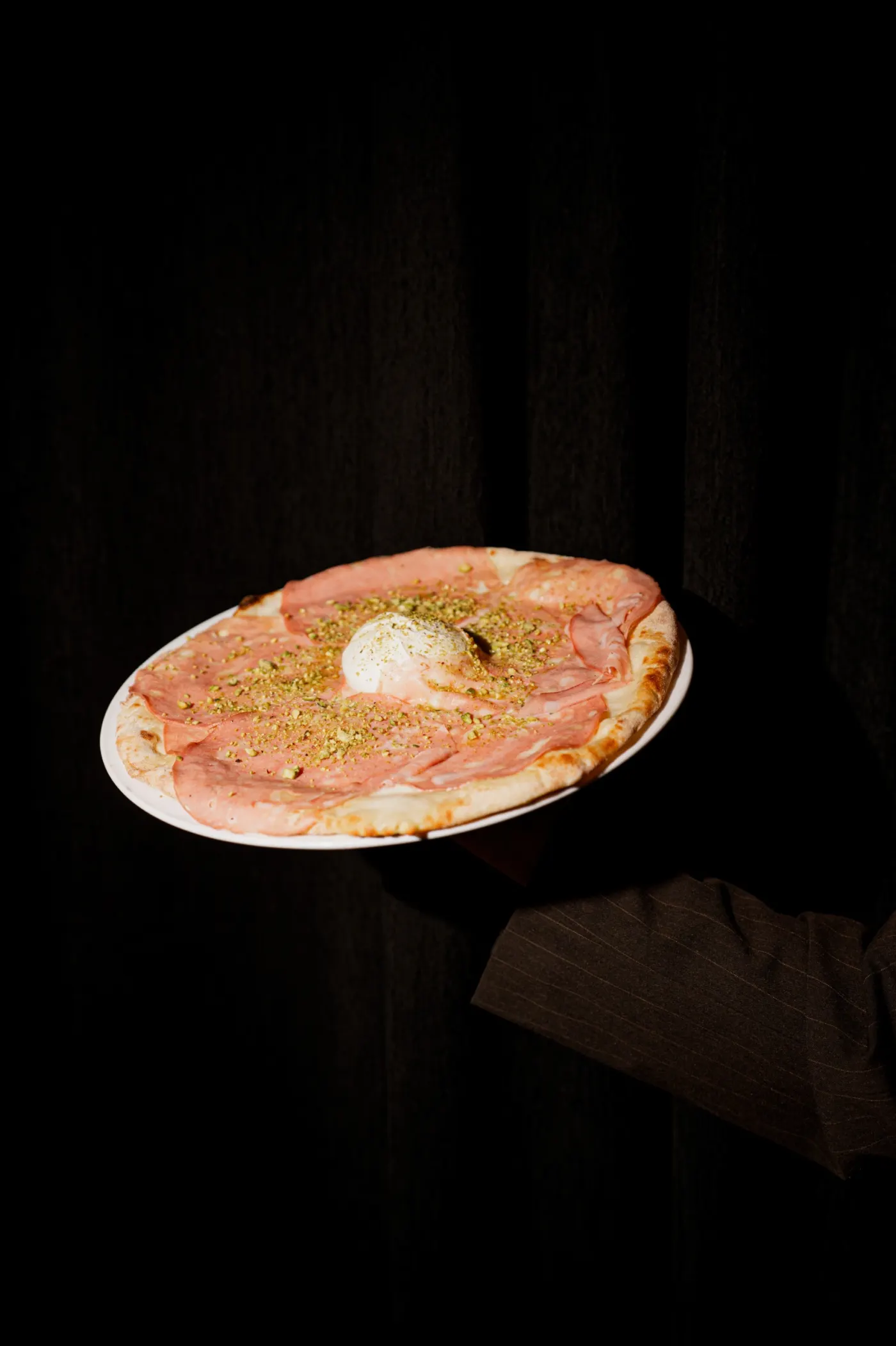 Una pizza con mortadela, pistachos picados y una bola de burrata, sostenida sobre un fondo oscuro.